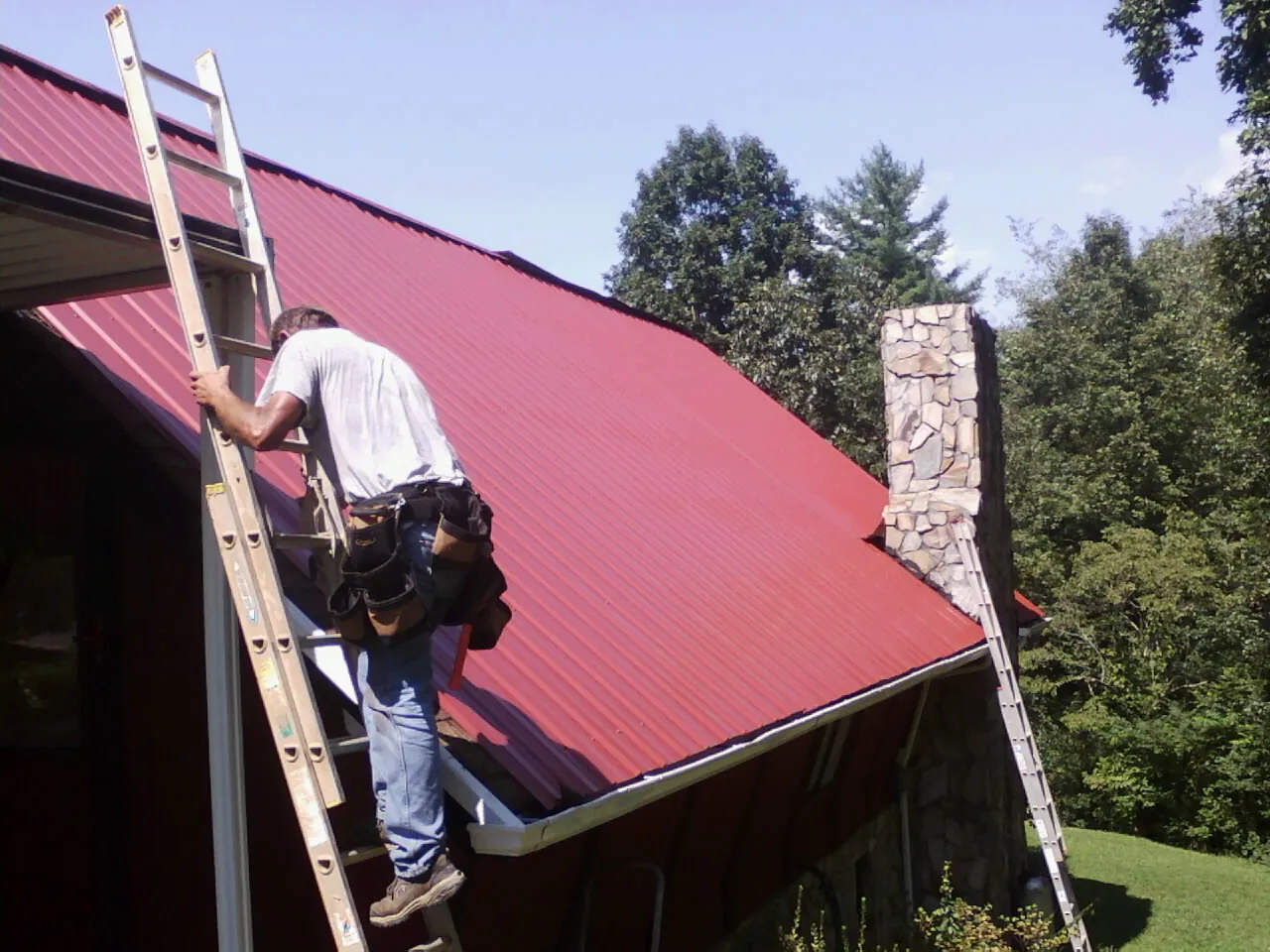 Roofer on a ladder beside a red metal roof and stone chimney — Soffit & Fascia Repair by Roofing Brothers in Boyers